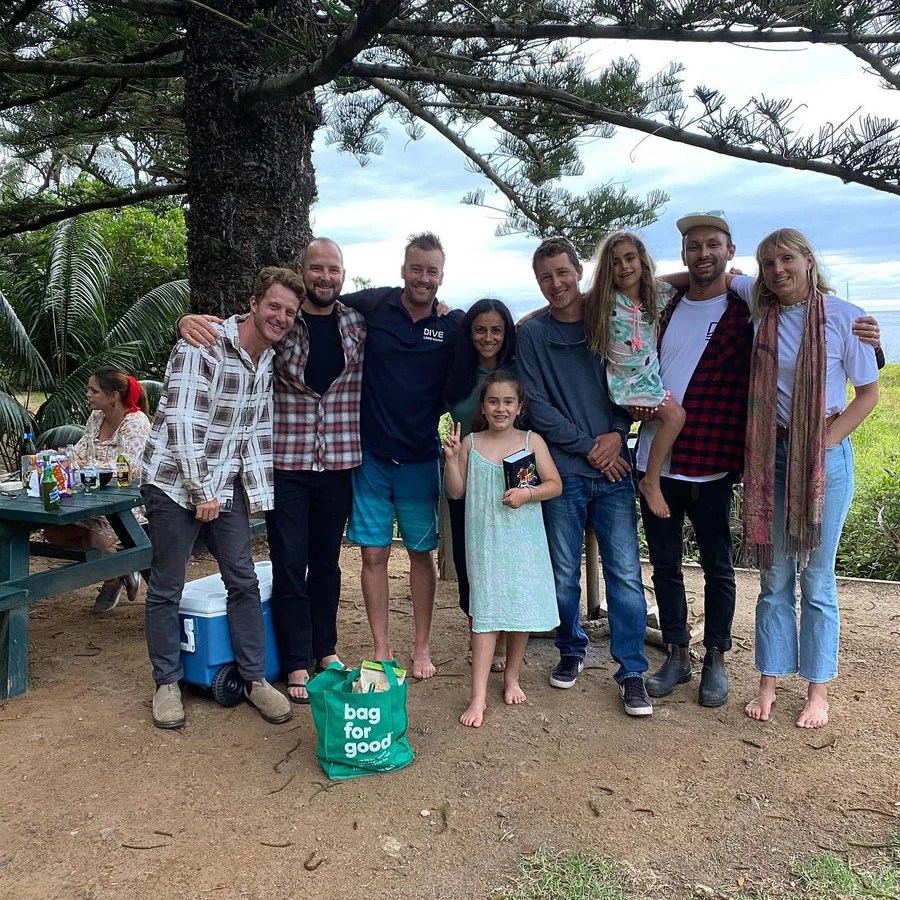 Group photo of eight adults and two children outdoors near a tree, with picnic items on a table.