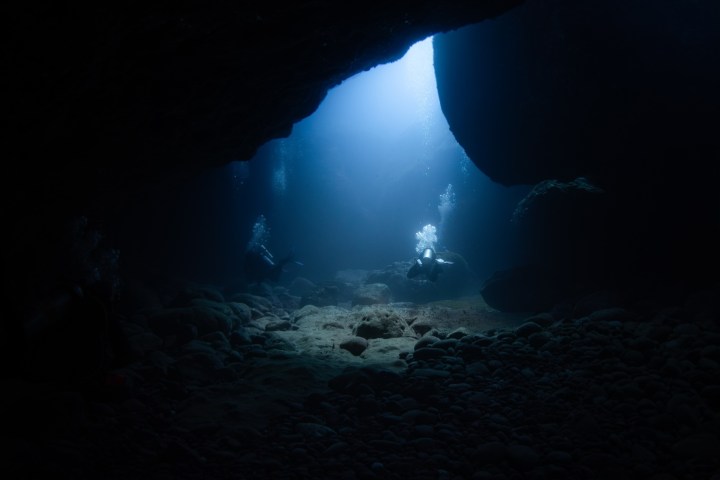 Two divers exploring an underwater cave with light streaming through the entrance.