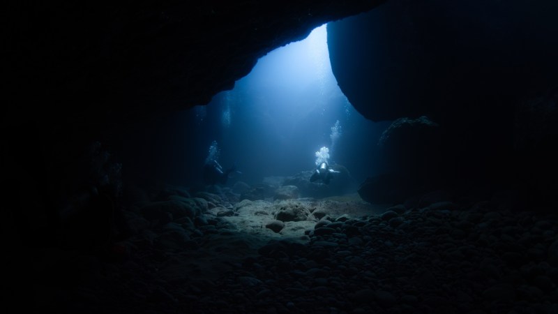 Two divers exploring an underwater cave with light streaming through the entrance.