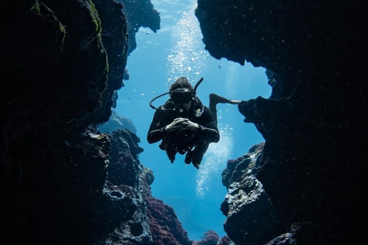 Scuba diver swimming through underwater rock arch, surrounded by blue water.