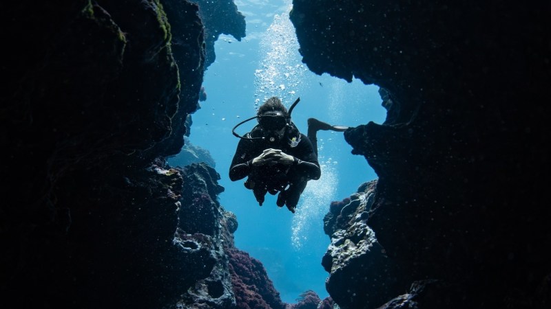 Scuba diver swimming through underwater rock arch, surrounded by blue water.