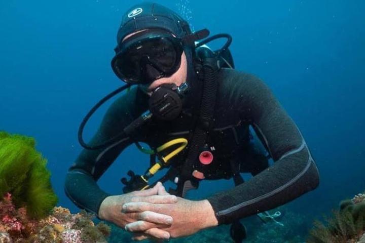 Scuba diver in black suit underwater near coral reef, hands clasped.