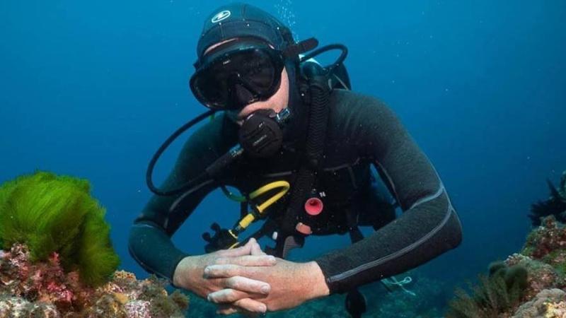 Scuba diver in black suit underwater near coral reef, hands clasped.