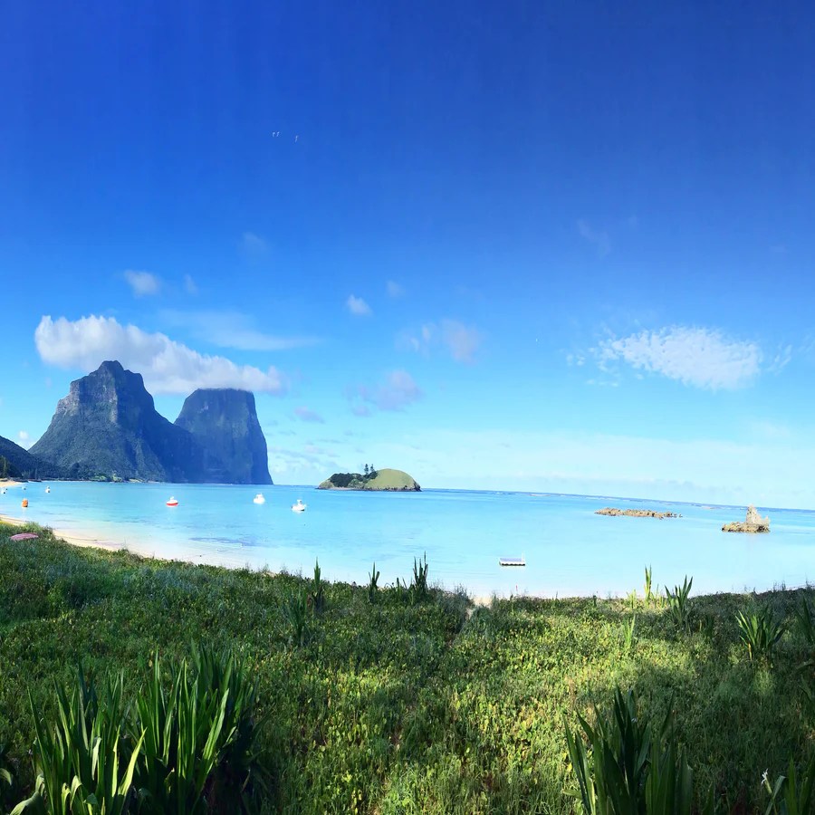 Tropical beach with blue water, green vegetation, and distant mountains under clear skies.
