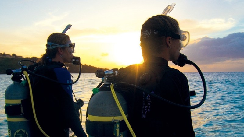 Two scuba divers in wetsuits watch sunset over ocean.