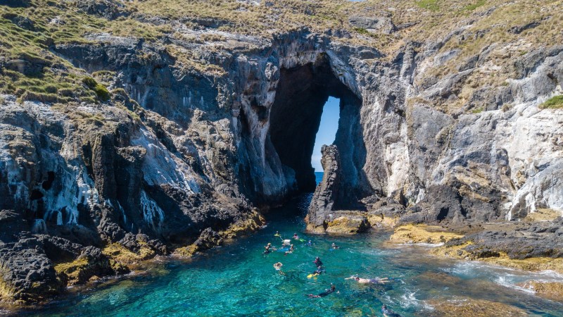 Snorkelers in clear water near a large sea cave surrounded by rocky cliffs.