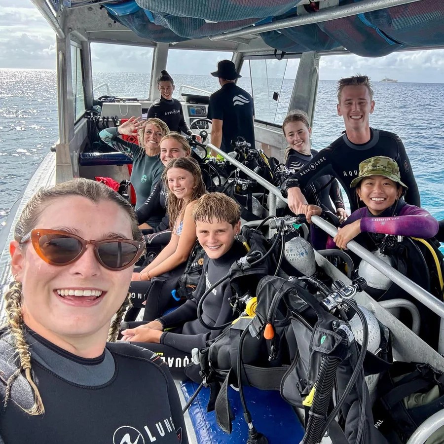 Group of people on a boat ready for scuba diving, wearing wetsuits and surrounded by diving gear.