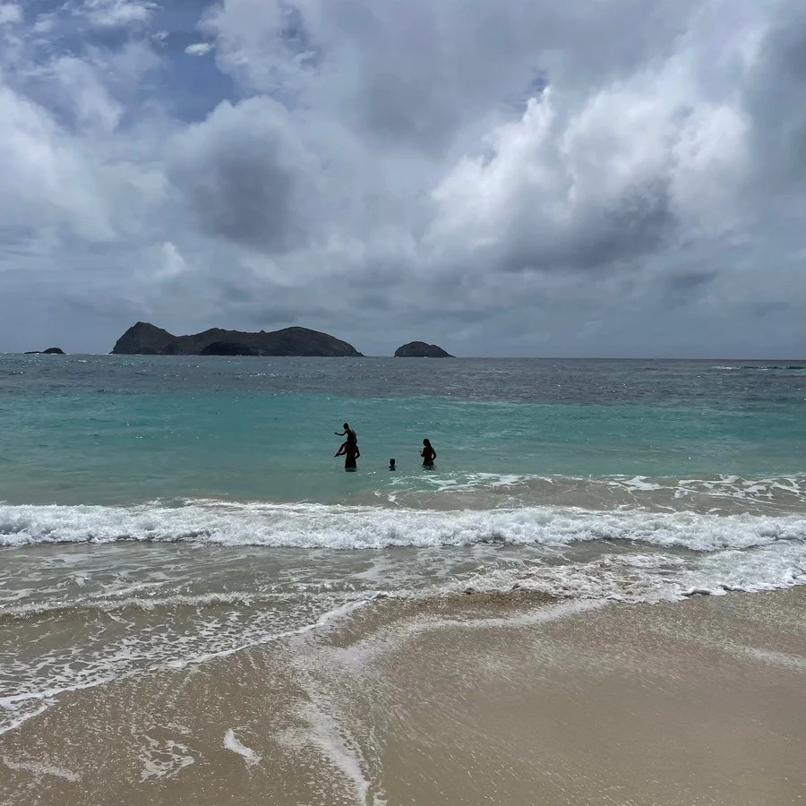 Three people swimming in the ocean near a sandy beach with cloudy sky.