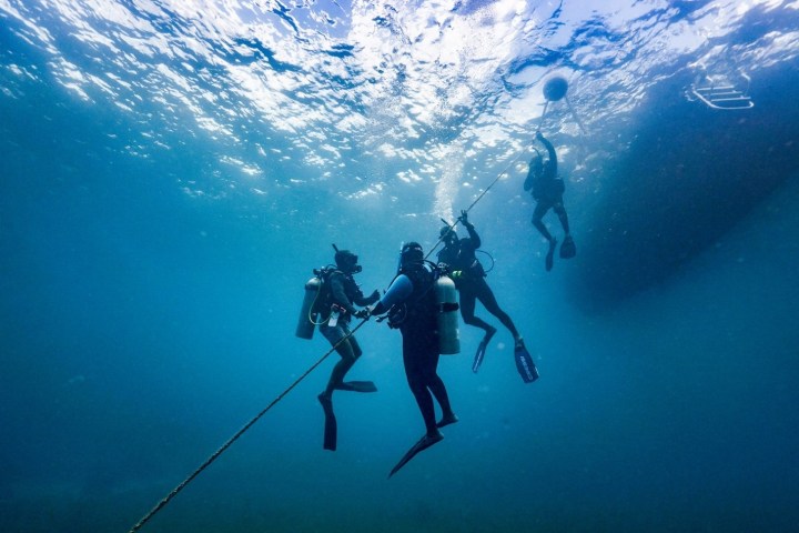 Four divers underwater near a rope leading to a buoy at the water surface.