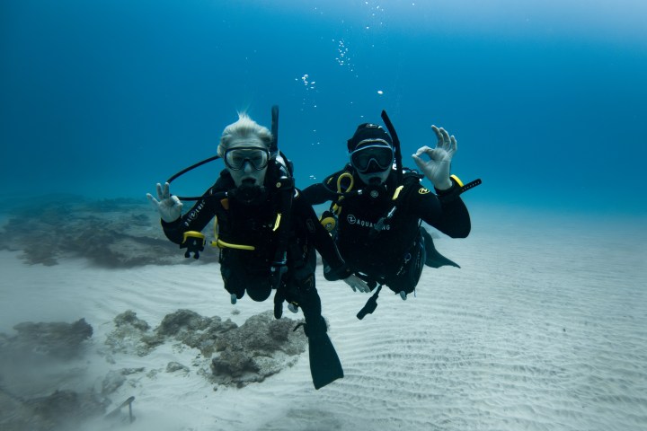 Two scuba divers underwater showing OK sign with clear ocean and sandy bottom.