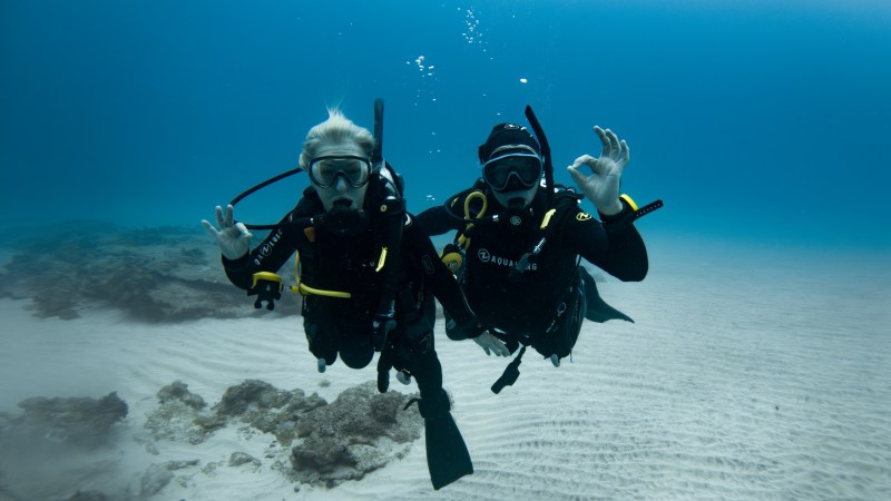 Two scuba divers underwater showing OK sign with clear ocean and sandy bottom.