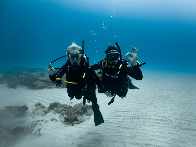 Two scuba divers underwater showing OK sign with clear ocean and sandy bottom.