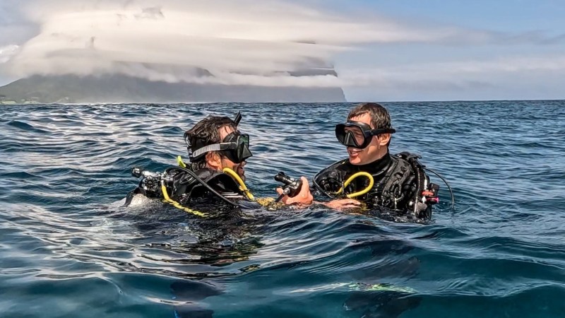 Two scuba divers in the ocean, with a cloud-covered mountain in the background.