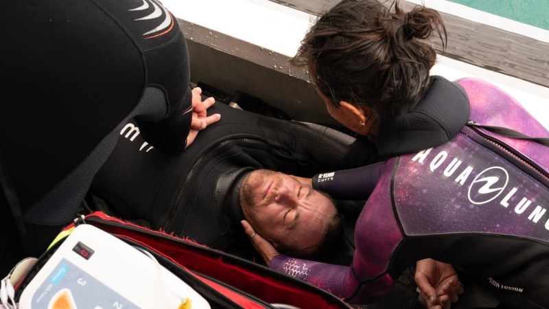Two divers in wetsuits assist an injured person lying on a boat.