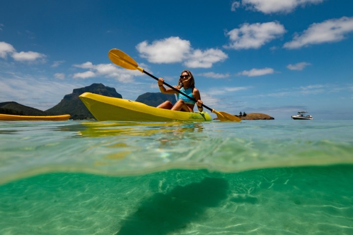 Person kayaking in clear water with mountains and a boat in the background under sunny skies.