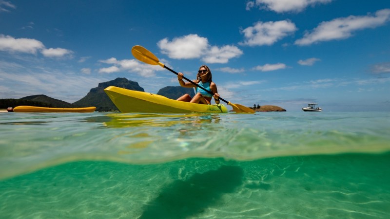 Person kayaking in clear water with mountains and a boat in the background under sunny skies.