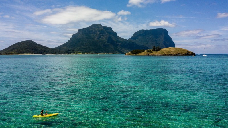 Person kayaking on clear turquoise water with mountains and islands in the background.