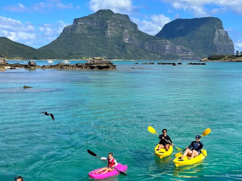 Three people kayaking in turquoise water with mountains in the background and a bird flying overhead.