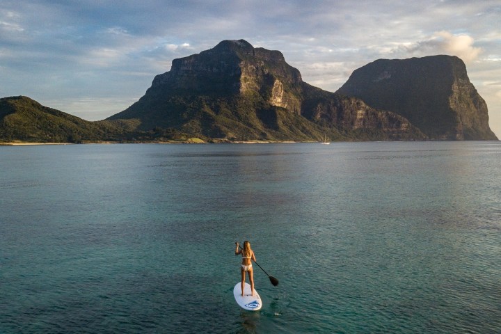 Person paddleboarding on calm water with large mountains in the background under a cloudy sky.
