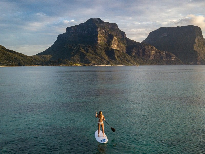 Person paddleboarding on calm water with large mountains in the background under a cloudy sky.