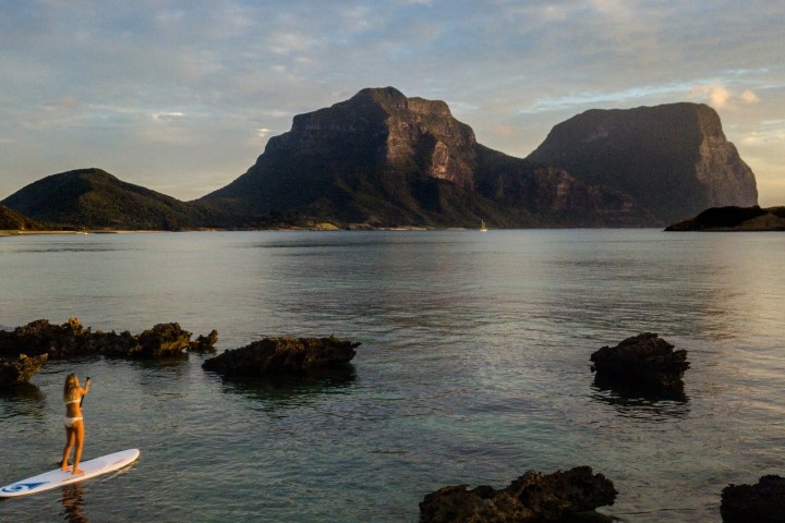 Person paddleboarding on calm water with mountains in the background at sunset.