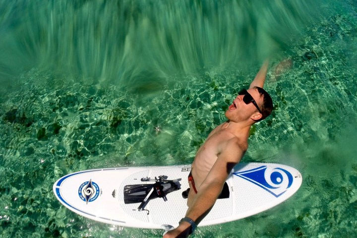 Man on paddleboard in clear water, wearing sunglasses and raising one arm.