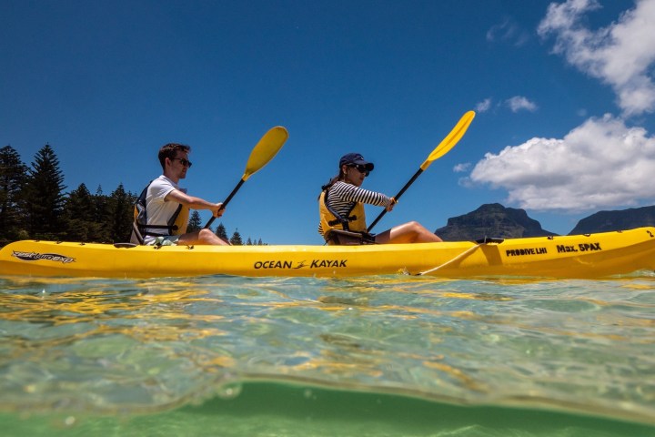 Two people kayaking in a yellow kayak on clear water with mountains and trees in the background.