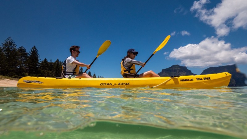 Two people kayaking in a yellow kayak on clear water with mountains and trees in the background.