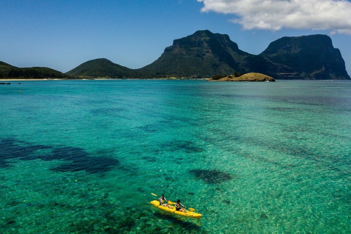 Two people kayaking on turquoise water with mountains in the background under a clear blue sky.