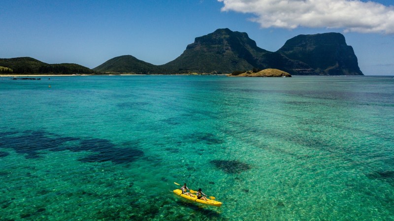 Two people kayaking on turquoise water with mountains in the background under a clear blue sky.