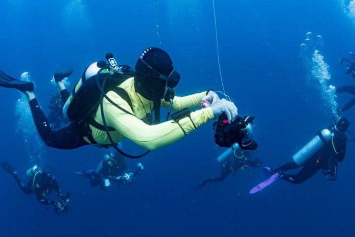 Group of scuba divers underwater, one holding a camera, surrounded by blue water and bubbles.