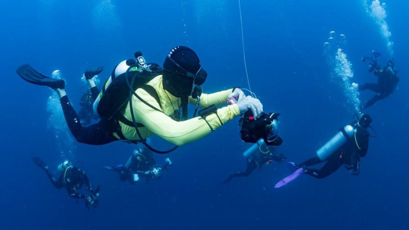 Group of scuba divers underwater, one holding a camera, surrounded by blue water and bubbles.
