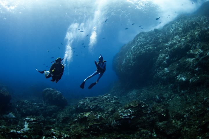 Two scuba divers swimming near rocks and fish in a deep blue ocean.