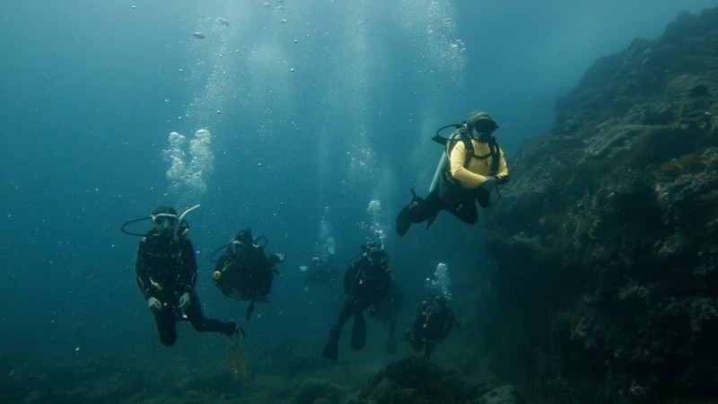 Scuba divers exploring underwater near a rocky reef, with bubbles rising to the surface.