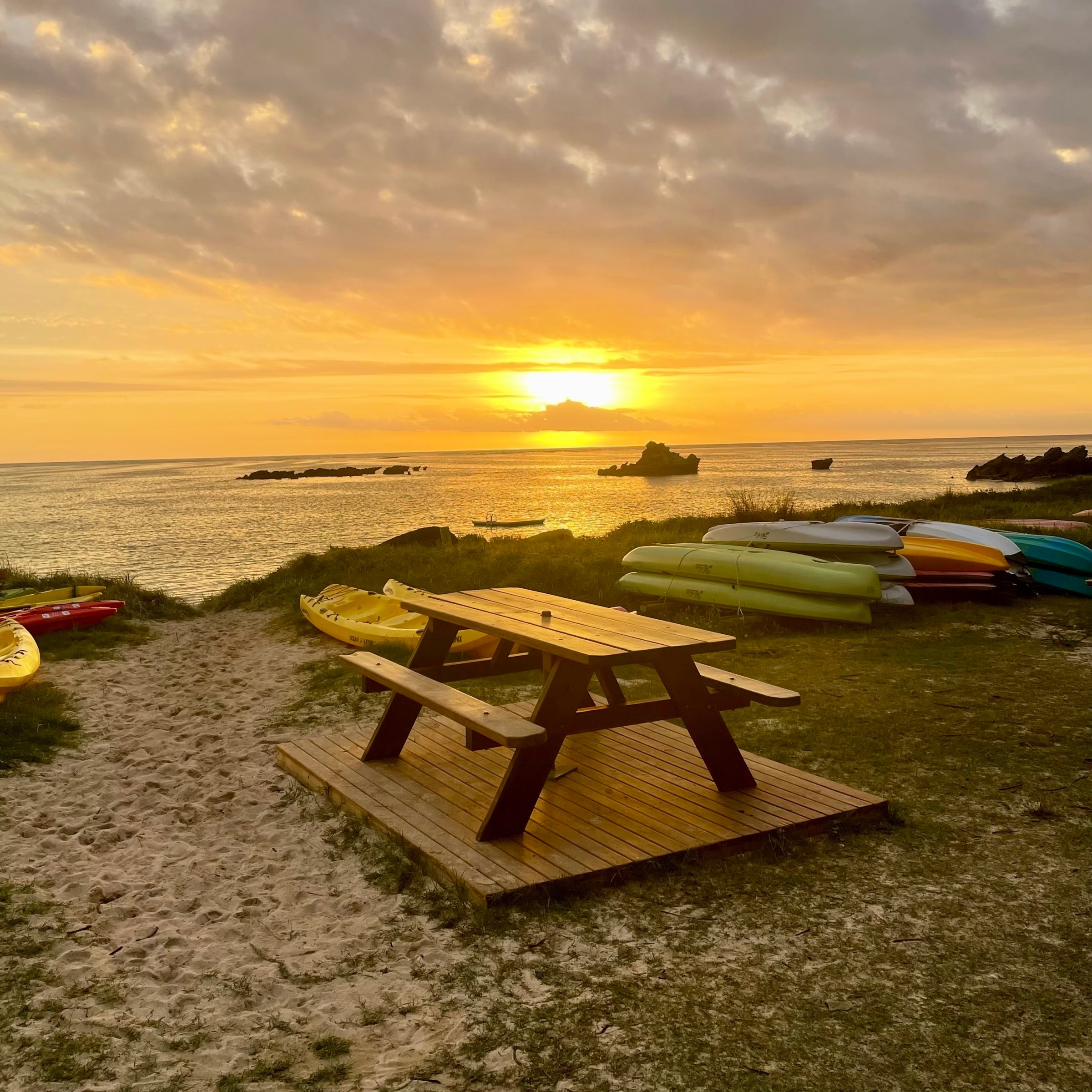 Sunset over a beach with picnic table and stacked kayaks.